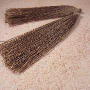May include: Two brown tassels with a silky, textured appearance. The tassels are made of numerous strands of thread, creating a flowing, decorative element. The tassels are likely used for embellishing clothing, accessories, or home decor items.