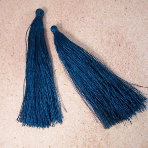 May include: Two navy blue tassels with a silky texture are displayed against a neutral background. The tassels are made of numerous fine threads, gathered at the top and flowing downwards. The color is a deep, rich blue.