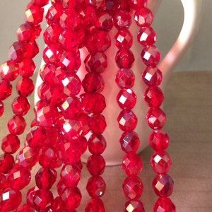 May include: Strands of faceted, bright red beads cascade from a white pitcher. The beads have a sparkling, gem-like quality, reflecting light. The beads are likely for jewelry making or other craft projects.