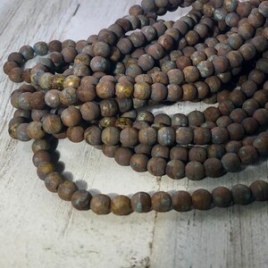 May include: Strands of round, rustic-looking beads in varying shades of brown with hints of blue and yellow. The beads have a textured, weathered appearance, suggesting an aged or antique aesthetic. The beads are likely used for jewelry making or crafting.