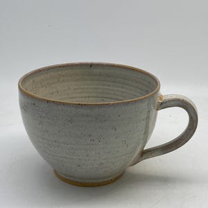 Può includere: Un'ampia tazza in ceramica maculata con smalto grigio chiaro e una visibile trama a coste orizzontali sull'esterno. Il bordo e il manico presentano un caldo tono bruno-aranciato, mentre la base mostra un'argilla color arancio-marrone.