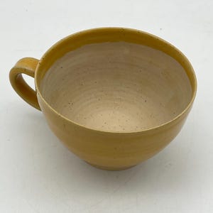 Può includere: Una tazza in ceramica fatta a mano con un corpo arrotondato e un manico singolo. La tazza ha un esterno giallo e un interno beige chiaro macchiettato. La tazza è progettata per contenere bevande calde come caffè o tè.