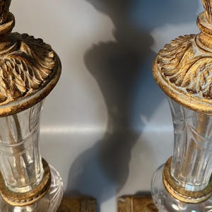 May include: Two decorative table lamps featuring clear glass bases and gold-coloured, detailed tops. The lamps have a classic, vintage design, with a decorative, leaf-like pattern on the top. The lamps are lit, casting shadows on the background.