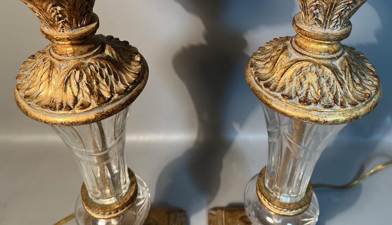 May include: Two decorative table lamps featuring clear glass bases and gold-coloured, detailed tops. The lamps have a classic, vintage design, with a decorative, leaf-like pattern on the top. The lamps are lit, casting shadows on the background.