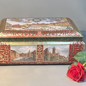 May include: Rectangular metal box with a hinged lid. The box is red with gold accents and features illustrations of a city scene. A red rose and green leaves are in the foreground.
