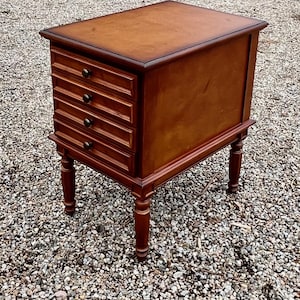 May include: A wooden side table with a warm brown finish. It features four drawers on one side with brass knobs and rests on four turned legs. The table is approximately 24 inches tall and 20 inches wide.
