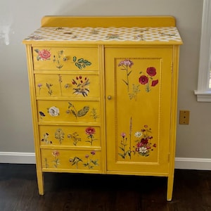 May include: A bright yellow wooden cabinet featuring five stacked drawers on the left and a single door on the right. The surface is decorated with colorful floral decals, and the top flat surface displays a white and yellow diamond pattern.