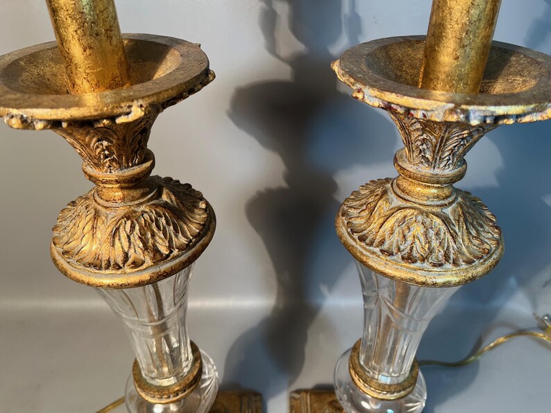 May include: Two ornate gold-coloured candle holders with clear glass bases. The candle holders have a detailed, vintage design with a fluted glass body and a decorative, gold-coloured top.