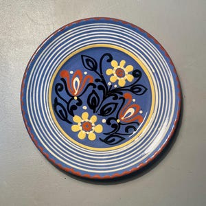 May include: A round, blue ceramic plate with a floral design. The plate features a blue background with white stripes around the edge. The centre has a yellow ring with a floral pattern in red, yellow, and black.