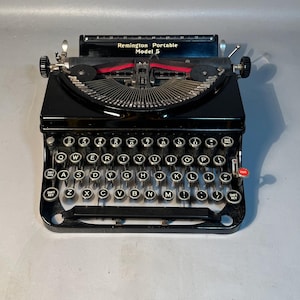 May include: A black Remington Portable Model 5 typewriter. The typewriter has a QWERTY keyboard with white keys and black lettering. The ribbon is red. The text "Remington Portable Model 5" is visible on the top of the typewriter.