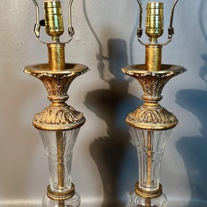 May include: Two ornate table lamps with gold bases and accents. They have clear glass columns and gold-toned metal lamp holders. The lampshades are not included. The lamps are designed with a classic, vintage aesthetic. The height of the lamps is approximately 60 cm.