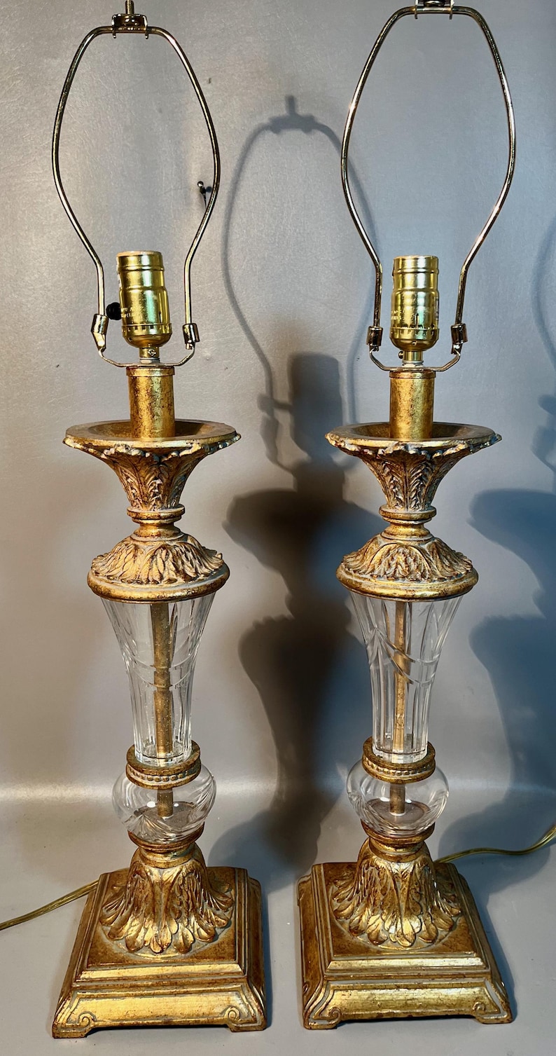 May include: Two ornate table lamps with gold bases and accents. They have clear glass columns and gold-toned metal lamp holders. The lampshades are not included. The lamps are designed with a classic, vintage aesthetic. The height of the lamps is approximately 60 cm.