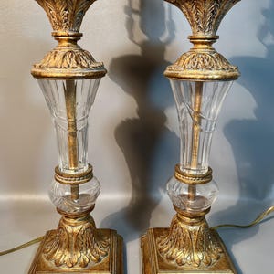 May include: Two decorative table lamps with gold-coloured bases and accents. Each lamp has a clear, fluted glass centre and a leaf-like design. The lamps have a classic, vintage aesthetic, suitable for a living room.