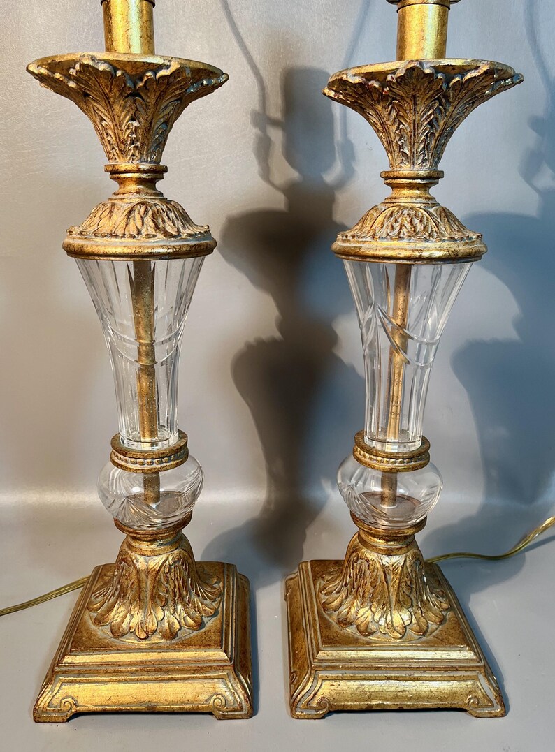 May include: Two decorative table lamps with gold-coloured bases and accents. Each lamp has a clear, fluted glass centre and a leaf-like design. The lamps have a classic, vintage aesthetic, suitable for a living room.