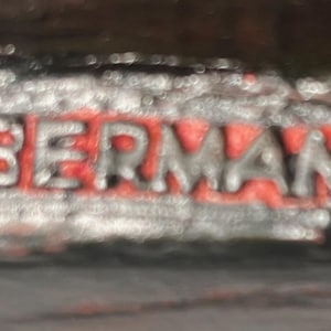 May include: Close-up of a metal object with the word "BERMAN" in red letters. The word is inside a silver-coloured, textured frame. The copyright symbol is visible before the word "BERMAN".