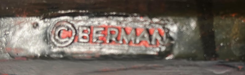 May include: Close-up of a metal object with the word "BERMAN" in red letters. The word is inside a silver-coloured, textured frame. The copyright symbol is visible before the word "BERMAN".