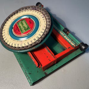 May include: A vintage Simplex typewriter toy in green, red, and blue. The circular dial features letters and numbers, with the brand name prominently displayed. The toy has a rectangular base and a roller for paper.