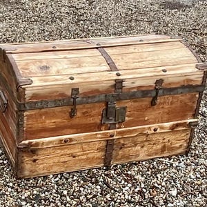 May include: A vintage wooden trunk with a curved top and metal hardware. The trunk is made of light brown wood with darker brown metal straps and corner protectors. The trunk has a metal lock and handle. The trunk is sitting on a bed of small pebbles.
