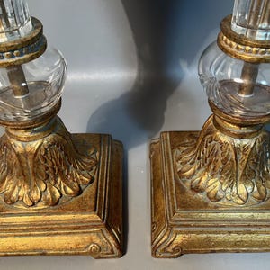 May include: A pair of antique table lamps featuring a gold-coloured, ornate base and clear glass accents. The base has a square shape with a detailed leaf design. The lamps have a clear glass globe and a clear, ridged glass column.