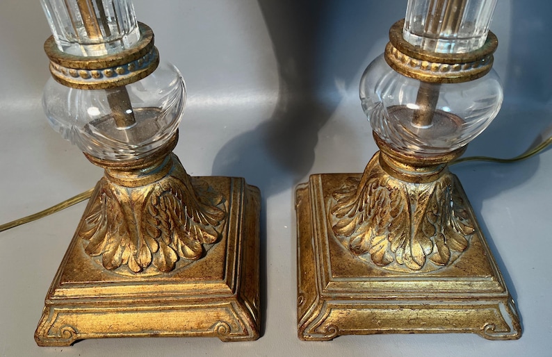 May include: A pair of antique table lamps featuring a gold-coloured, ornate base and clear glass accents. The base has a square shape with a detailed leaf design. The lamps have a clear glass globe and a clear, ridged glass column.
