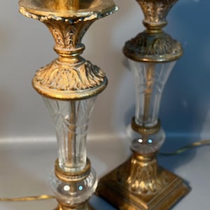 May include: A pair of vintage table lamps with gold-toned metal bases and accents. The lamps have clear glass columns and square bases. The lampshades are not visible. The lamps are connected to electrical cords.