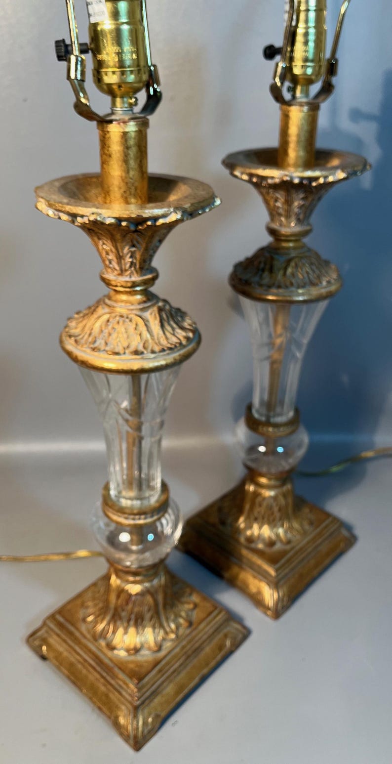 May include: A pair of vintage table lamps with gold-toned metal bases and accents. The lamps have clear glass columns and square bases. The lampshades are not visible. The lamps are connected to electrical cords.