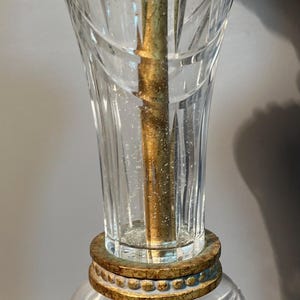 May include: Close-up of a vintage lamp with a clear, etched glass shade and a gold-coloured metal base. The lamp features a decorative, ornate design and a central brass rod.
