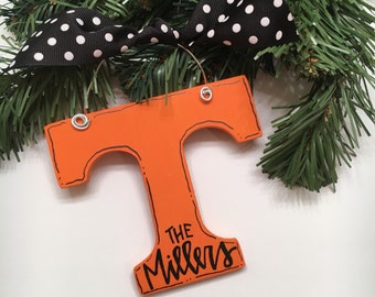 May include: Orange wooden letter "T" Christmas ornament with the text "The Millers" in black script. The ornament is trimmed in black and has silver eyelets for hanging. A black ribbon with white polka dots is attached to the ornament.