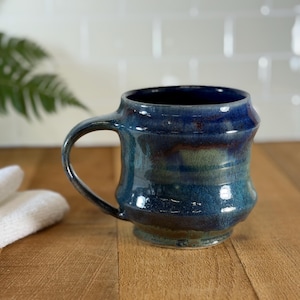 Op de afbeelding: Een handgemaakte keramische mok met een donkerblauwe glazuur, met een afgeronde body en een stevige handgreep. De mok heeft een getextureerd oppervlak en een unieke vorm, perfect voor koffie of thee. De mok is ongeveer 10 cm hoog.