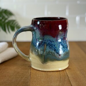 Puede incluir: Una taza de cerámica hecha a mano con un esmalte único. La taza presenta un interior rojo oscuro, que pasa a tonos azules y verdes en el exterior, con una base beige y un asa curva. La taza está diseñada para bebidas calientes.
