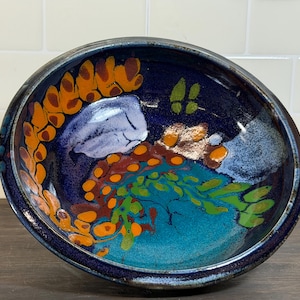May include: A handmade ceramic bowl with a dark blue exterior and a vibrant interior design. The interior features orange, green, and blue abstract patterns. The bowl is a unique piece of art pottery.