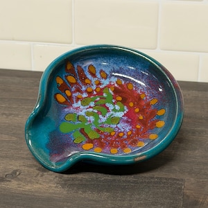 Op de afbeelding: Een keramische lepelsteun met een turquoise buitenkant en een kleurrijk interieurontwerp. De binnenkant heeft een patroon van rode, oranje, groene en gele stippen en wervelingen. De lepelsteun staat op een donker houten oppervlak.