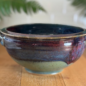 May include: A handmade ceramic bowl with a dark blue interior and a gradient exterior of red, blue, and green hues. The bowl has two small handles and sits on a wooden surface. The bowl is suitable for serving food.