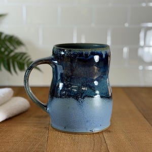 Op de afbeelding: Een handgemaakte keramische mok met een grote handgreep. De mok heeft een verloop van blauwtinten, van een donker marineblauw aan de bovenkant naar een lichter blauw aan de basis. De mok staat op een houten oppervlak.