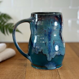 May include: A handmade ceramic mug with a large handle. The mug has a glossy finish with shades of teal, blue, and dark brown. The mug is sitting on a wooden surface.
