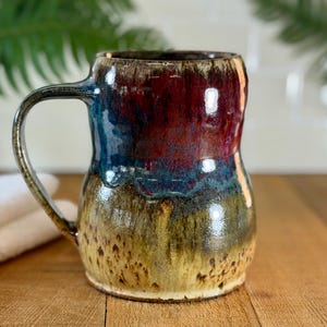 May include: A handmade ceramic mug with a large handle. The mug features a glossy finish with a blend of colors, including deep red, blue, and yellow-brown. The mug is approximately 5 inches tall.