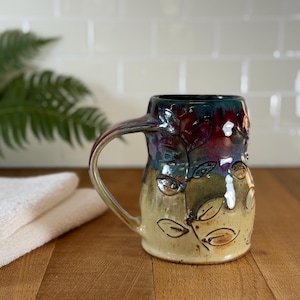May include: A handmade ceramic mug with a leaf design. The mug features a color gradient from a light yellow base to a dark blue rim, with red and green accents. The handle is a light yellow color. The mug is approximately 5 inches tall.