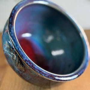 Può includere: Una ciotola in ceramica fatta a mano con una finitura lucida e iridescente. La ciotola presenta una miscela di tonalità blu, verde acqua e rosse, con un bordo strutturato e un interno liscio. Il design della ciotola è sia funzionale che decorativo.