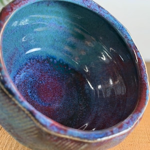 Op de afbeelding: Een handgemaakte keramische kom met een diepe, afgeronde vorm. De kom heeft een glanzend, veelkleurig glazuur in tinten blauw, blauwgroen en bordeaux. De rand heeft een roodbruin accent en de buitenkant heeft een gestructureerd, geribbeld ontwerp.
