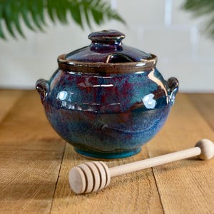Puede incluir: Un tarro de miel de cerámica vidriada en tonos azul, turquesa y burdeos, con tapa a juego. Un cucharón de miel de madera clara descansa junto al tarro. El tarro tiene dos asas pequeñas. El conjunto está sobre una superficie de madera.