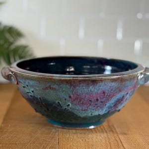 Op de afbeelding: Een handgemaakte keramische kom met een donkerblauwe binnenkant en een getextureerde buitenkant. De kom heeft een mix van teal, paarse en bruine tinten, met kleine handvatten aan weerszijden. Decoratieve bladerpatronen zijn zichtbaar.