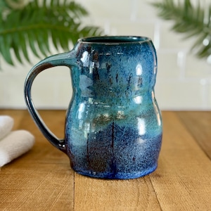 May include: A handmade ceramic mug with a unique shape and a large handle. The mug features a gradient of blue hues, transitioning from a deep navy at the base to lighter turquoise shades towards the top. The mug is approximately 5 inches tall.