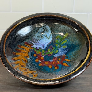 May include: A handmade ceramic bowl with a dark blue and brown rim. The interior features a colorful abstract design with orange, red, green, and blue hues. The bowl is approximately 10 inches in diameter.