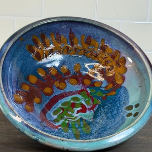 May include: A handmade ceramic bowl with a vibrant, abstract design. The bowl features a deep blue interior with accents of orange, red, and green. The rim is a lighter blue-green. This decorative bowl is ideal for serving or display.