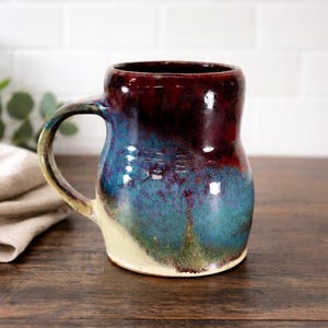 May include: A handmade ceramic mug with a unique color gradient. The top of the mug is a deep burgundy, transitioning to a vibrant blue and green towards the base. It has a curved handle and a cream-colored base.