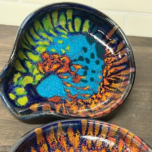 May include: A set of handmade ceramic spoon rests with a unique, colorful design. The interior features a vibrant mix of turquoise, orange, and green against a dark blue background. The spoon rests have an organic shape and glossy finish.