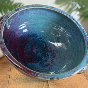 May include: A handmade ceramic bowl with a striking blend of blue, teal, and purple hues. The bowl features a glossy finish and a textured rim with rope-like handles. The interior showcases a marbled effect, adding depth to the design. A decorative piece for the home.