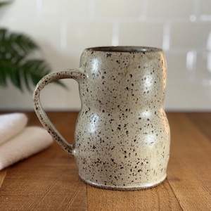 May include: A speckled ceramic mug with a unique hourglass shape and a curved handle. The mug is beige with dark brown speckles and a dark brown interior. It sits on a wooden surface, with a white towel and green plant in the background.