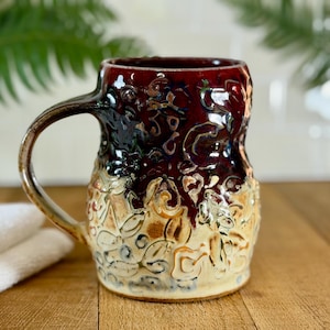 May include: A handcrafted ceramic mug with a dark red glazed interior and a textured, floral-patterned exterior. The mug has a brown handle and a beige base, perfect for enjoying coffee or tea.