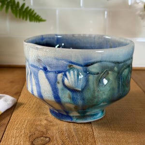 Op de afbeelding: Een keramische kom met een reactief glazuur in tinten blauw, groenblauw en crème. Het buitenoppervlak is versierd met reliëfmotieven van schelpen en verticale druippatronen, terwijl de binnenkant is afgewerkt met een glad, diepblauw glazuur.